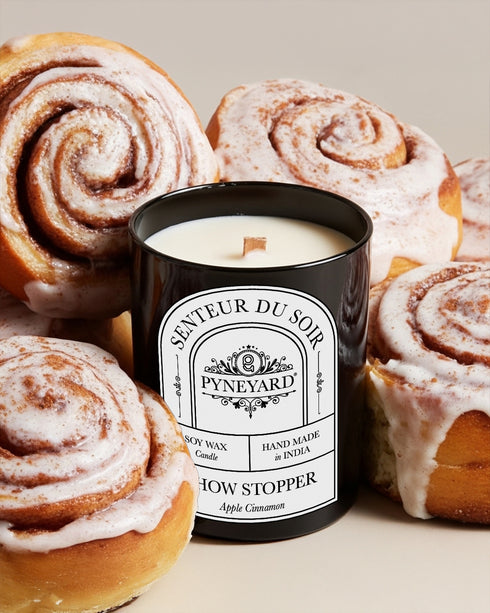 Showstopper Apple Cinnamon Candle labeled 'Pyneyard' surrounded by cinnamon rolls on a light background from the collection Reverie.