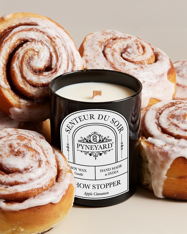 Showstopper Apple Cinnamon Candle labeled 'Pyneyard' surrounded by cinnamon rolls on a light background from the collection Reverie.