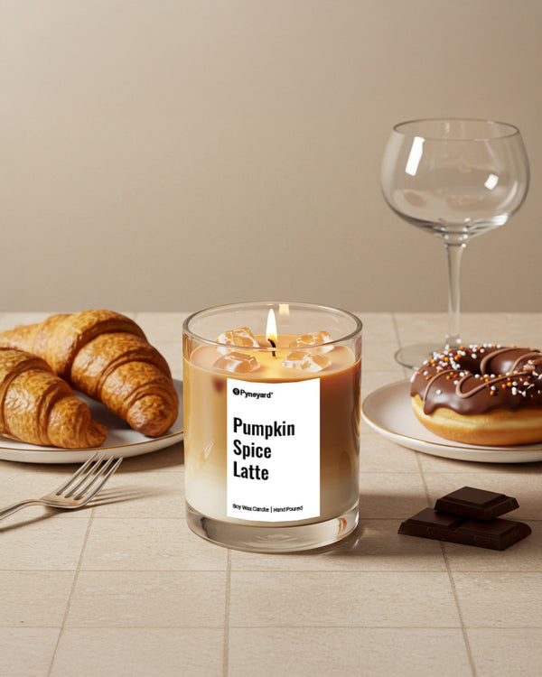 Pyneyard Candle labeled 'Pumpkin Spice Latte' on a table with croissants, a donut, and a glass.